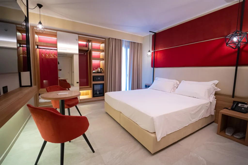 Pictured is a room with two single beds. The walls are red and white. In front of the beds is a round wooden table and two red velvet armchairs. On one side of the room is a large wardrobe with a minibar and a safe. To the side is a window door.
