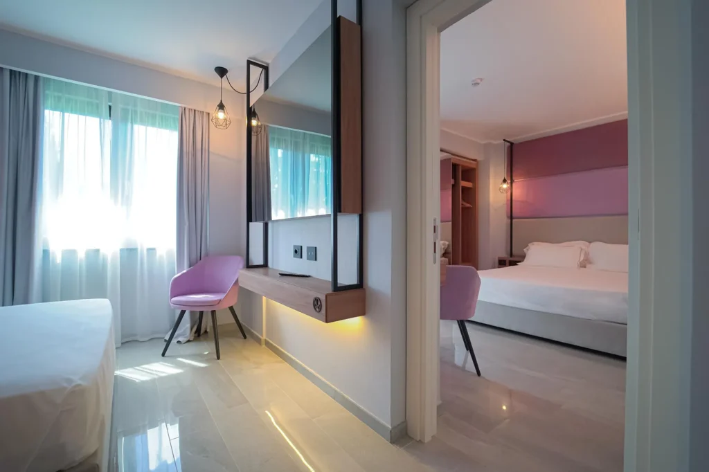 Pictured is a room with a double bed. The walls are white and pink. In front of the bed a small round wooden table and two pink velvet armchairs. The second room can also be glimpsed.