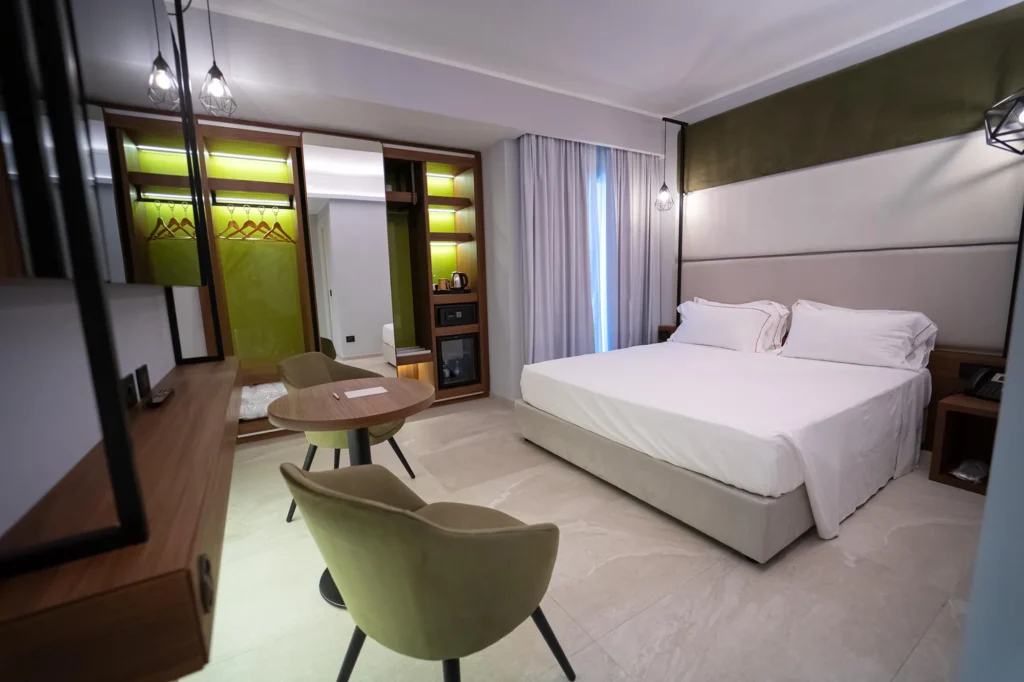 The picture shows a room with a double bed. The walls are white and green. In front of the beds is a round wooden table and two red velvet armchairs. On one side of the room is a large wardrobe with a mini-bar and a safe. To the side is a window door.