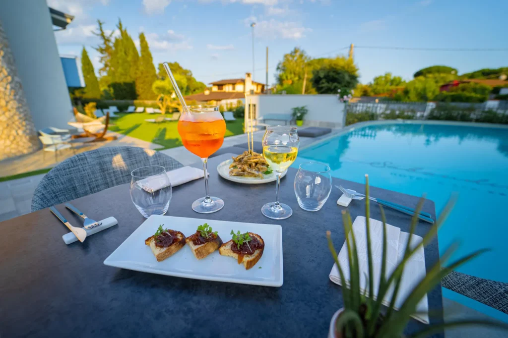 Nell'immagine un tavolino del ristorante a bordo piscina. Sul tavolino: un calice di vino, uno spritz e due piatti di portata.